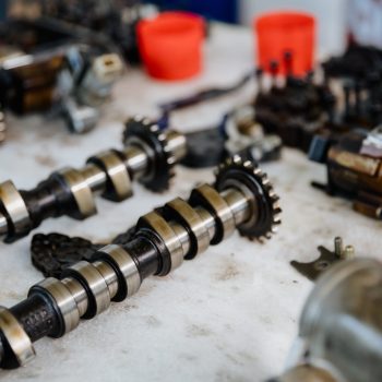 Auf einer Werkbank liegen mehrere Nockenwellen und weitere Motorteile, die zur Reparatur oder Wartung ausgebaut wurden. Die Bauteile sind aus Metall und zeigen Zahnräder, Lagerstellen und verschiedene Befestigungselemente. Im Hintergrund sind weitere Autoteile und Werkstattzubehör wie rote Becher zu sehen.
