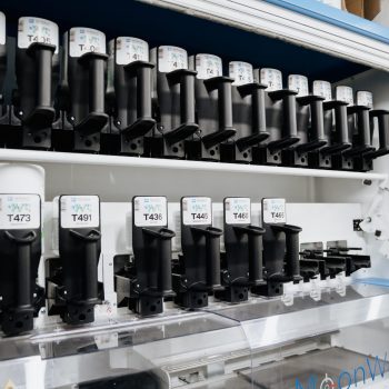 Das Bild zeigt eine Regalwand mit zahlreichen Farbdosierern, die ordentlich in zwei Reihen angeordnet sind. Jede Kartusche ist mit einem Etikett und einer individuellen Nummer wie „T471“ versehen.