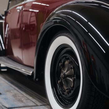 Detailaufnahme eines restaurierten Oldtimers mit glänzendem schwarz-rotem Lack und auffälligen Weißwandreifen. Zu sehen sind der hintere Kotflügel, die Radkappe und elegante Chromleisten. Das Fahrzeug steht auf einer Hebebühne in einer hellen Werkstattumgebung.