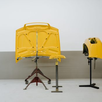 Das Bild zeigt drei frisch lackierte gelbe Karosserieteile, die auf speziellen Halterungen in einer Werkstatt stehen. Zu sehen sind eine Motorhaube, ein kleineres Verkleidungsteil und eine Stoßstange. Der Hintergrund ist schlicht und hebt die glänzenden Lackoberflächen deutlich hervor.