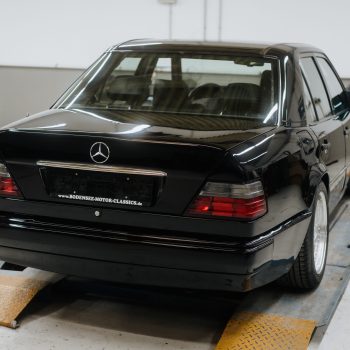 Das Bild zeigt einen schwarzen Mercedes-Benz Youngtimer auf einer Hebebühne in einer modernen Werkstatt. Der Wagen ist von hinten zu sehen, mit glänzendem Lack und klassischen Rückleuchten. Im Hintergrund steht ein weiteres Fahrzeug, die Umgebung wirkt aufgeräumt und technisch ausgestattet.