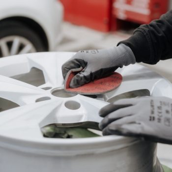 Eine Person schleift mit einem roten Schleifpad eine silberne Alufelge ab. Sie trägt graue Arbeitshandschuhe und bereitet die Felge sorgfältig für die weitere Bearbeitung vor. Im Hintergrund sind ein Auto und Werkstattausrüstung unscharf zu sehen.