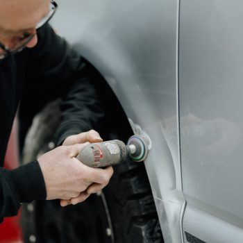 Eine Person bearbeitet mit einem elektrischen Schleifgerät den Radlauf eines Autos. Deutlich zu sehen sind die Handhabung des Werkzeugs und die bearbeitete Stelle im Lackbereich. Der Reifen und ein Teil des Kotflügels sind im Bildausschnitt erkennbar.