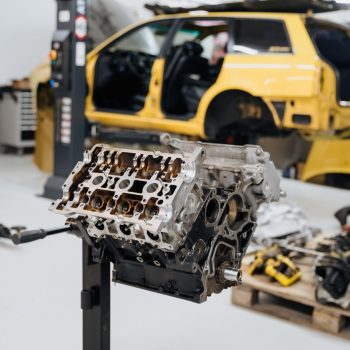 Im Vordergrund ist ein ausgebauter und teilweise zerlegter Motorblock auf einem Ständer zu sehen. Im Hintergrund steht ein gelbes Fahrzeug mit offenen Türen und ausgebauten Komponenten in einer Werkstatt. Auf einer Palette liegen weitere Motorteile und Werkzeuge bereit für Reparatur oder Montage.